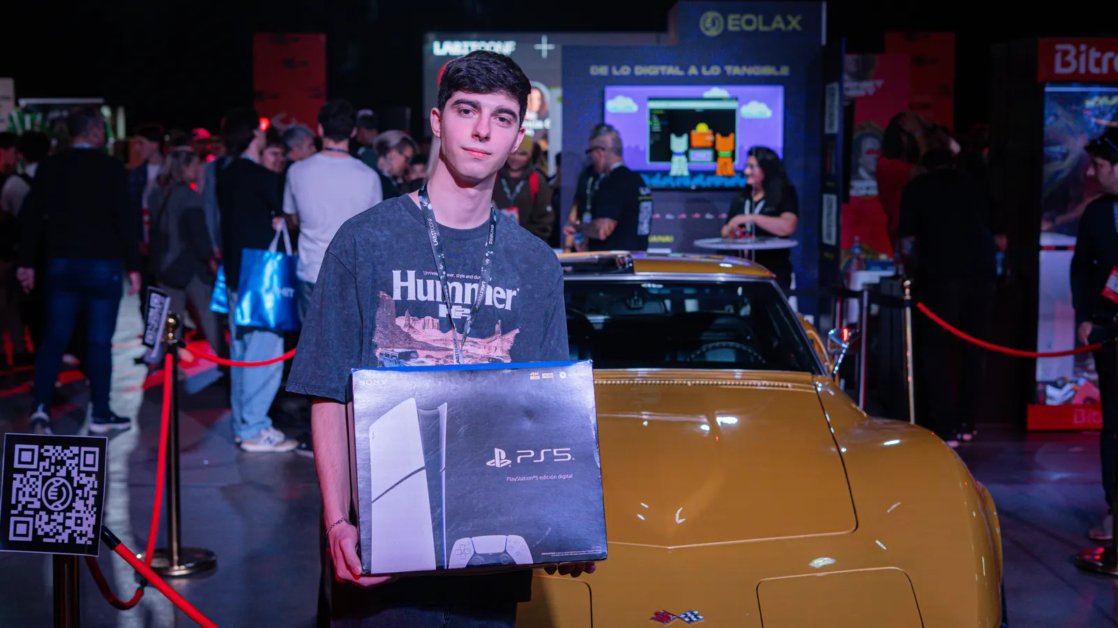 Facu sostiene la PlayStation 5 frente al Corvette Stingray dorado expuesto en el stand, con la conferencia de fondo.