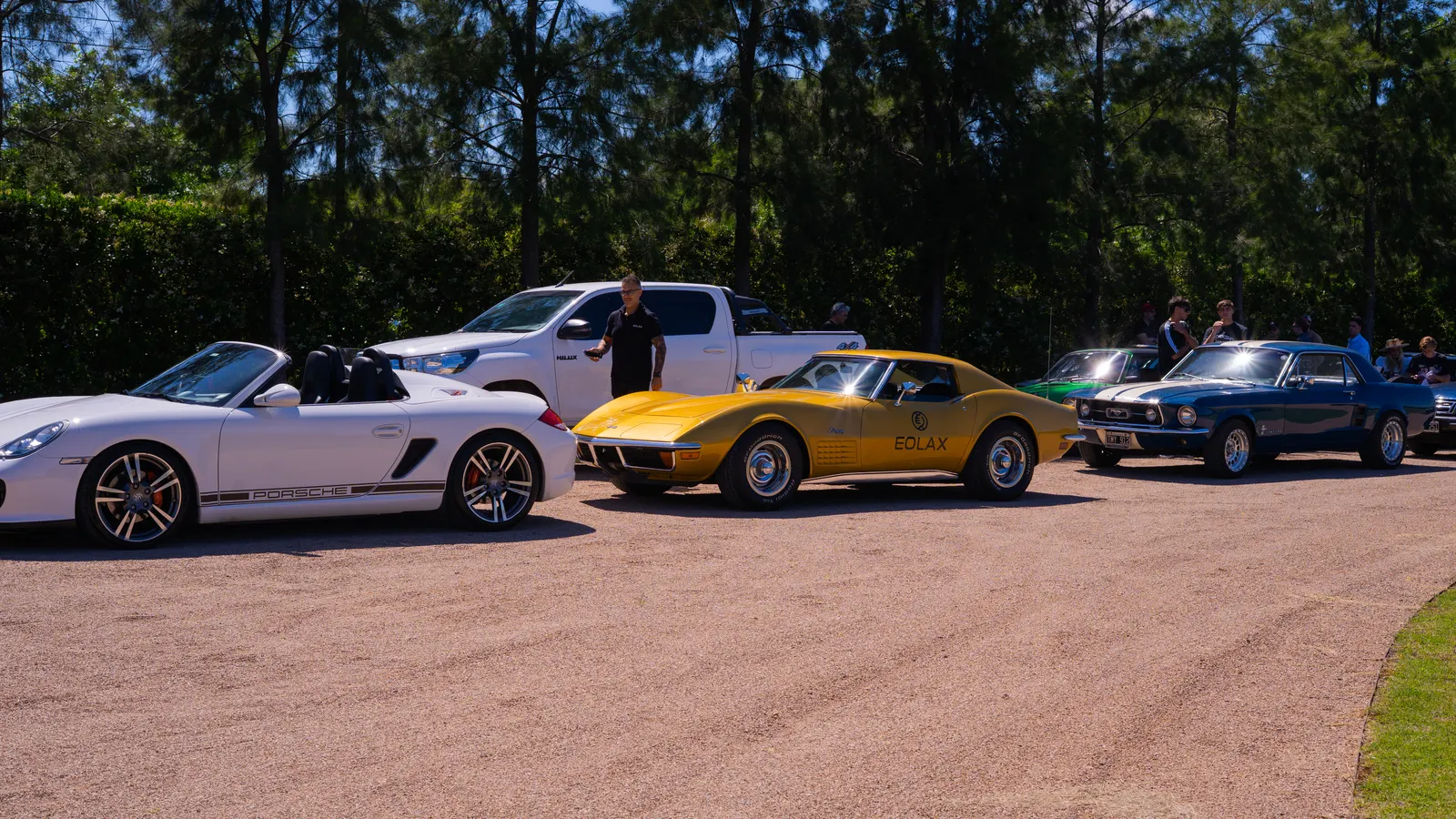 Autos clásicos estacionados sobre la grava en la largada: un descapotable blanco, el Corvette dorado y un cupé azul.