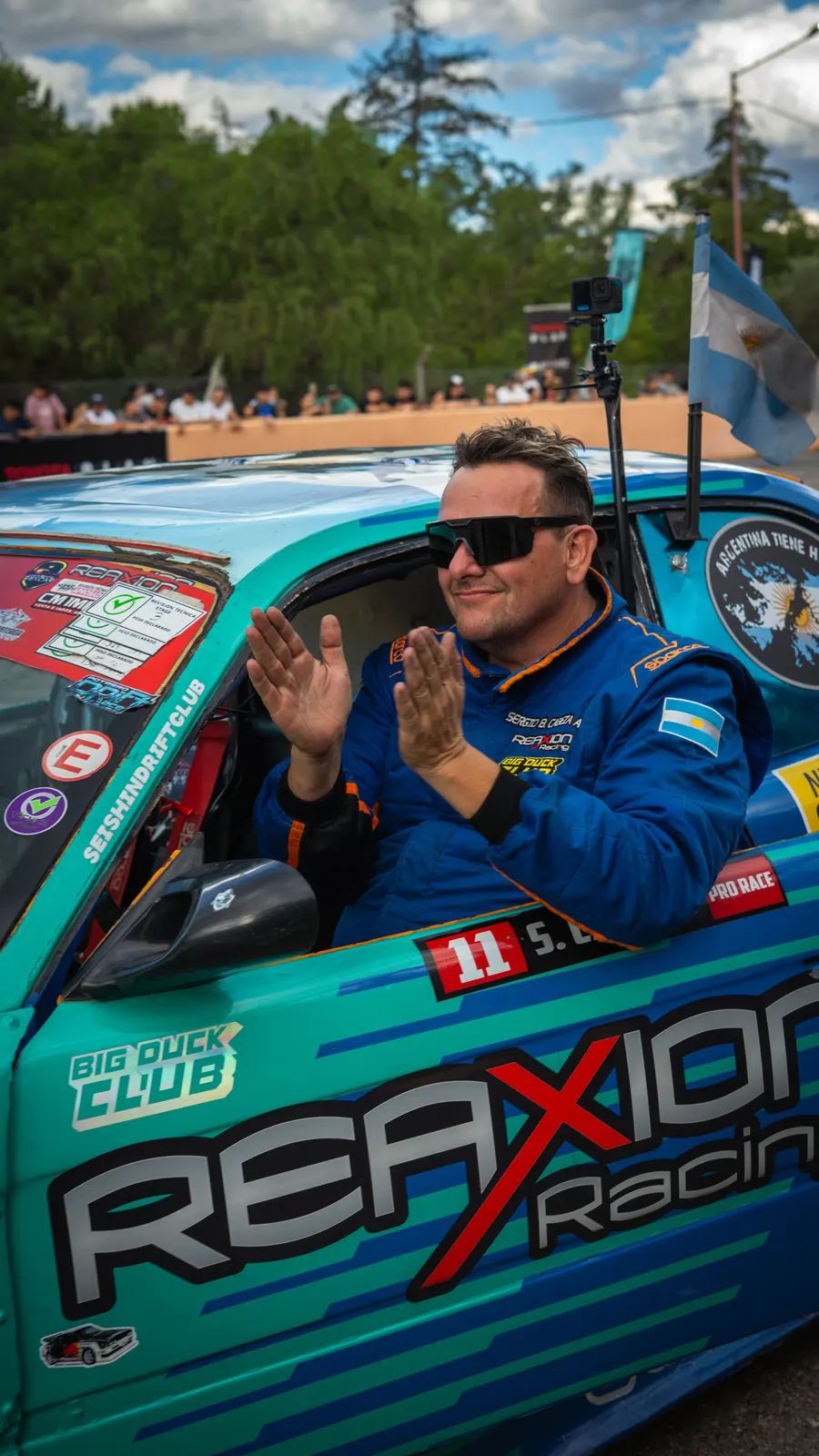 Un piloto saluda desde la ventanilla de su auto de drift turquesa, listo para salir a pista.