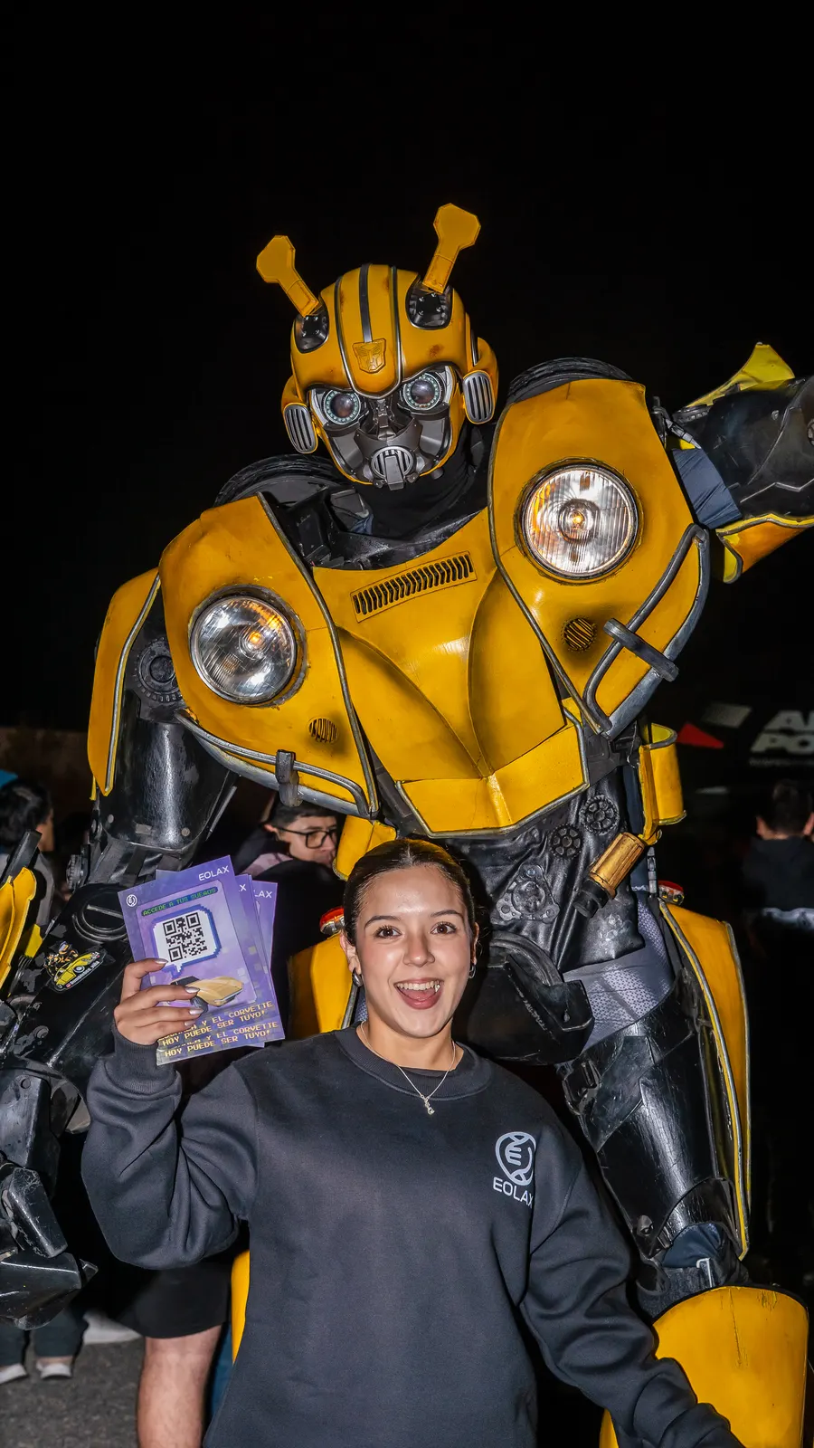 Una asistente muestra su ticket de Eolax frente a una figura gigante de Bumblebee, de noche.